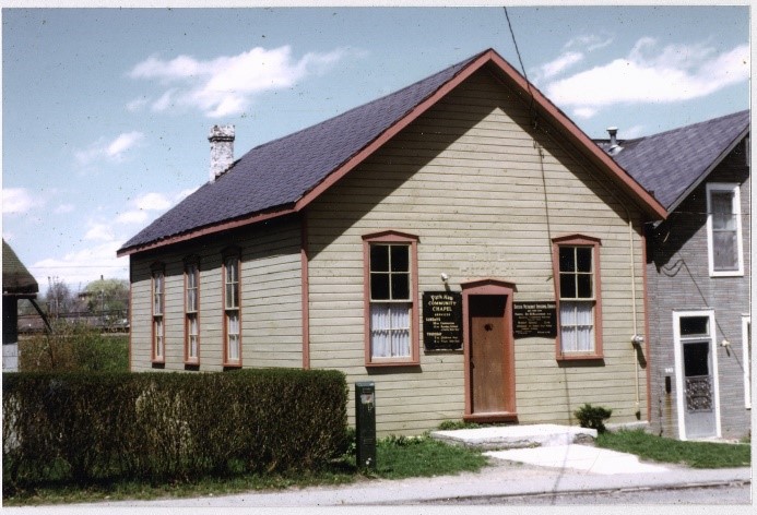 A colour photograph of the exterior of the Park Row Community Chapel in Woodstock, Ontario. The chapel is a wooden frame building with a sign next to the front door.