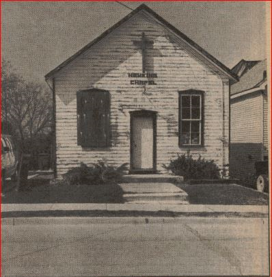 A newspaper photograph of the former Hawkins Chapel on Park Row in Woodstock, Ontario. The windows of the chapel are boarded up. A large wooden cross hangs above the front door.