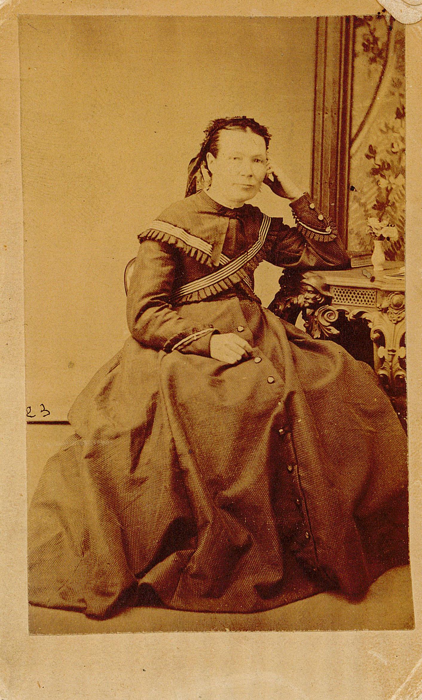 Studio portrait of Mary Marett, she is sitting in a chair with her head resting against her hand, her arm is leaning against an ornate table with a painting of flowers behind her.