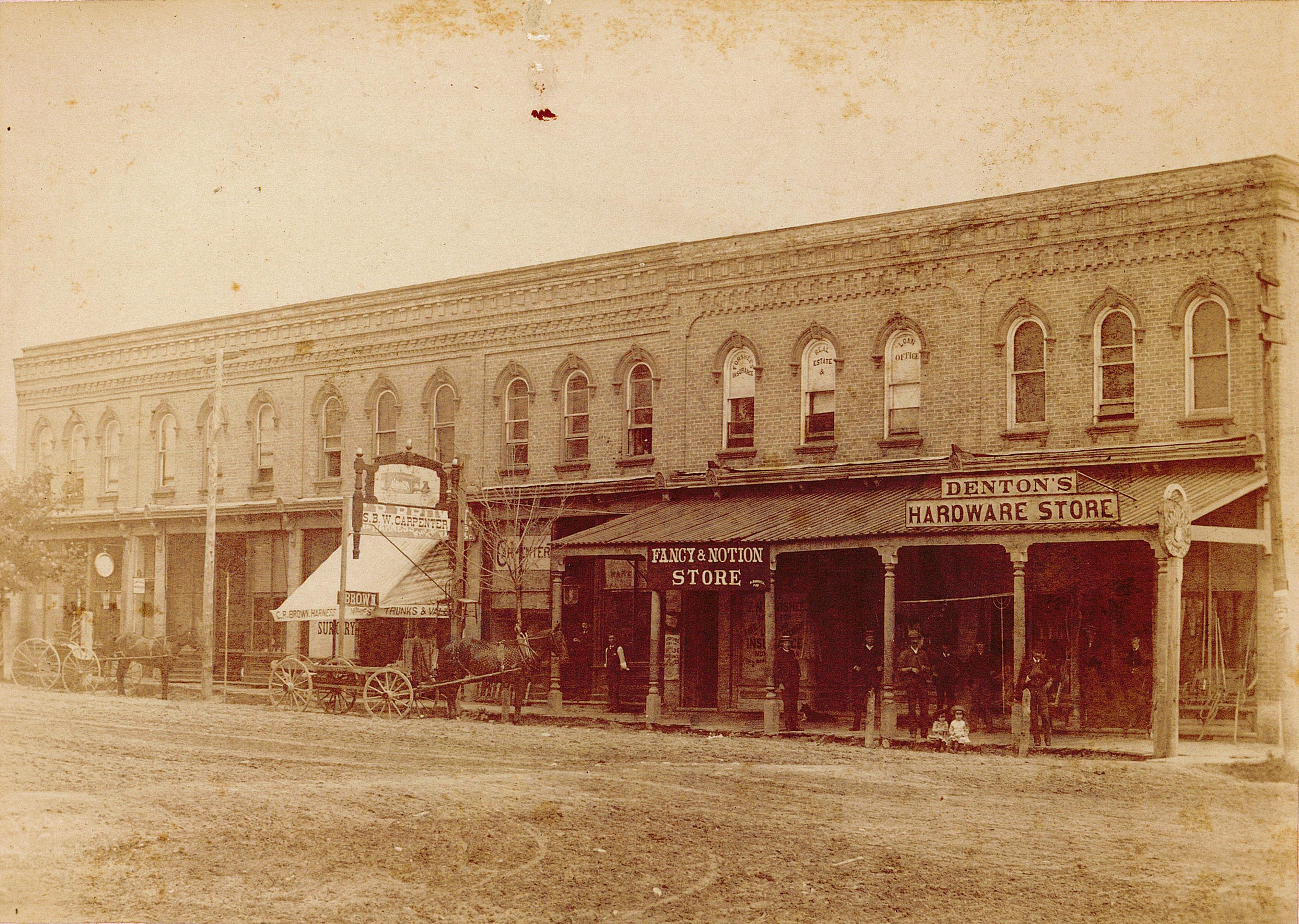 Streetscape of downtown Tillsonburg, horses and wagons are parked out front of stores. Store signs include S.B.W. Carpenter, Fancy & Notion Store, and Denton's Hardware.