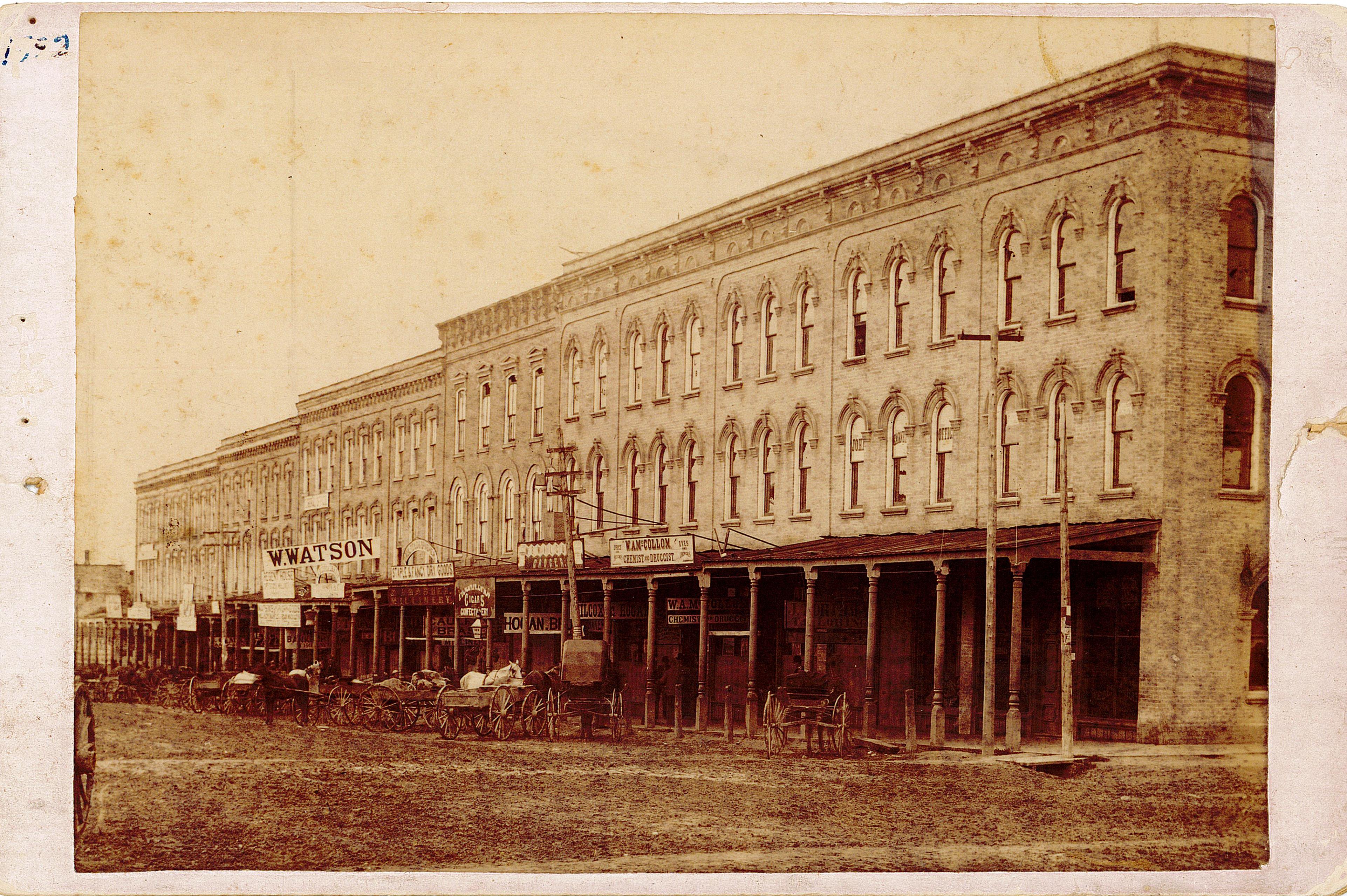 Brick buildings line a dirt road in Tillsonburg. Horses and wagons are parked at the front of the buildings. A sign for McCollom's drug store is on the front of one of the buildings.