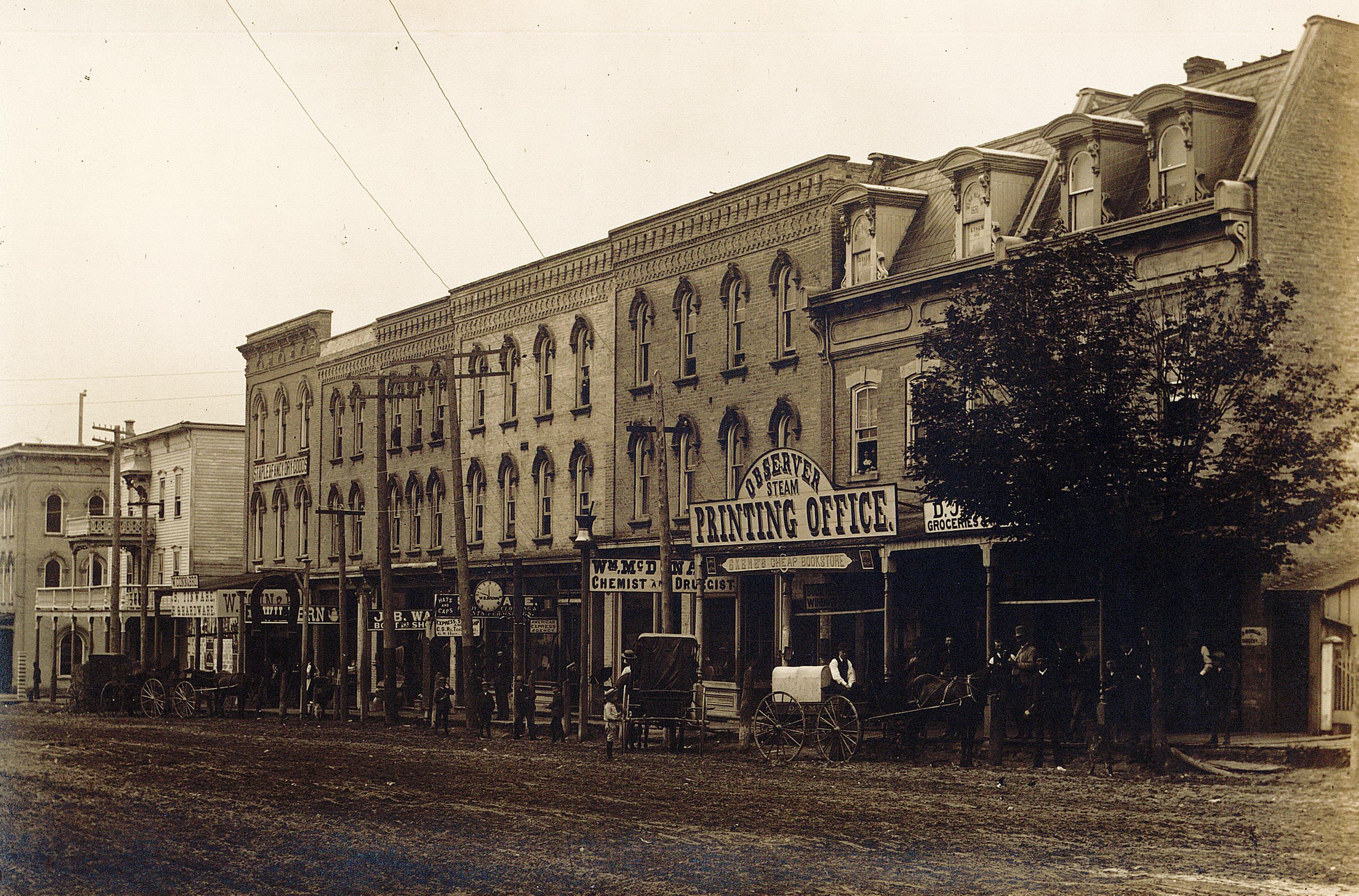 A streetscape of downtown Tillsonburg with a view of the Observer Printing Office. Buildings line the dirt road with store signs posted on the front of the buildings.