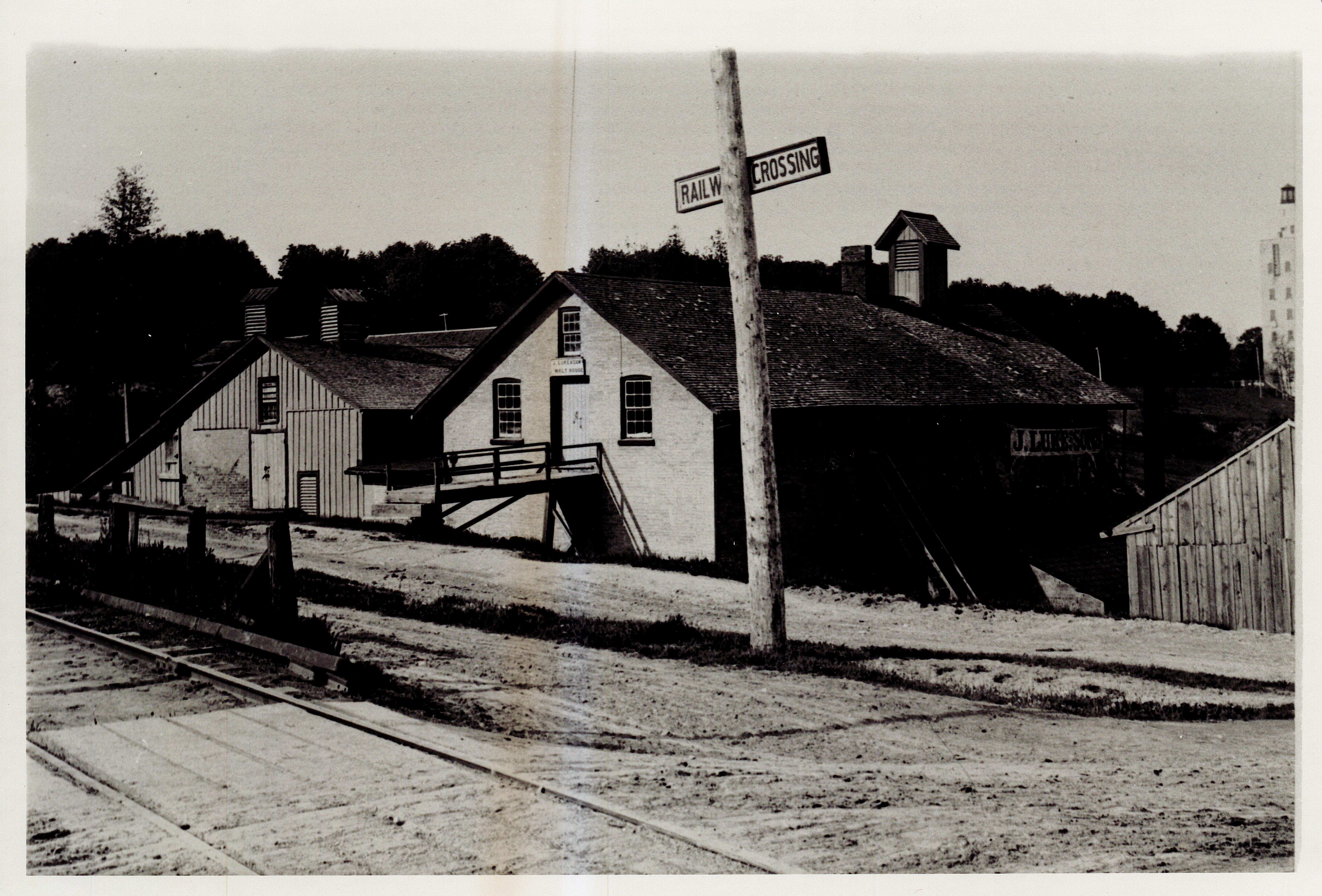 Two buildings, one brick and one wooden frame, are side by side. A railway line runs in front of the buildings. A wooden pole is off to the right with a sign posted to it that reads "railway crossing". The brick building has a sign above the door that reads: "Luke & Sons malt house".