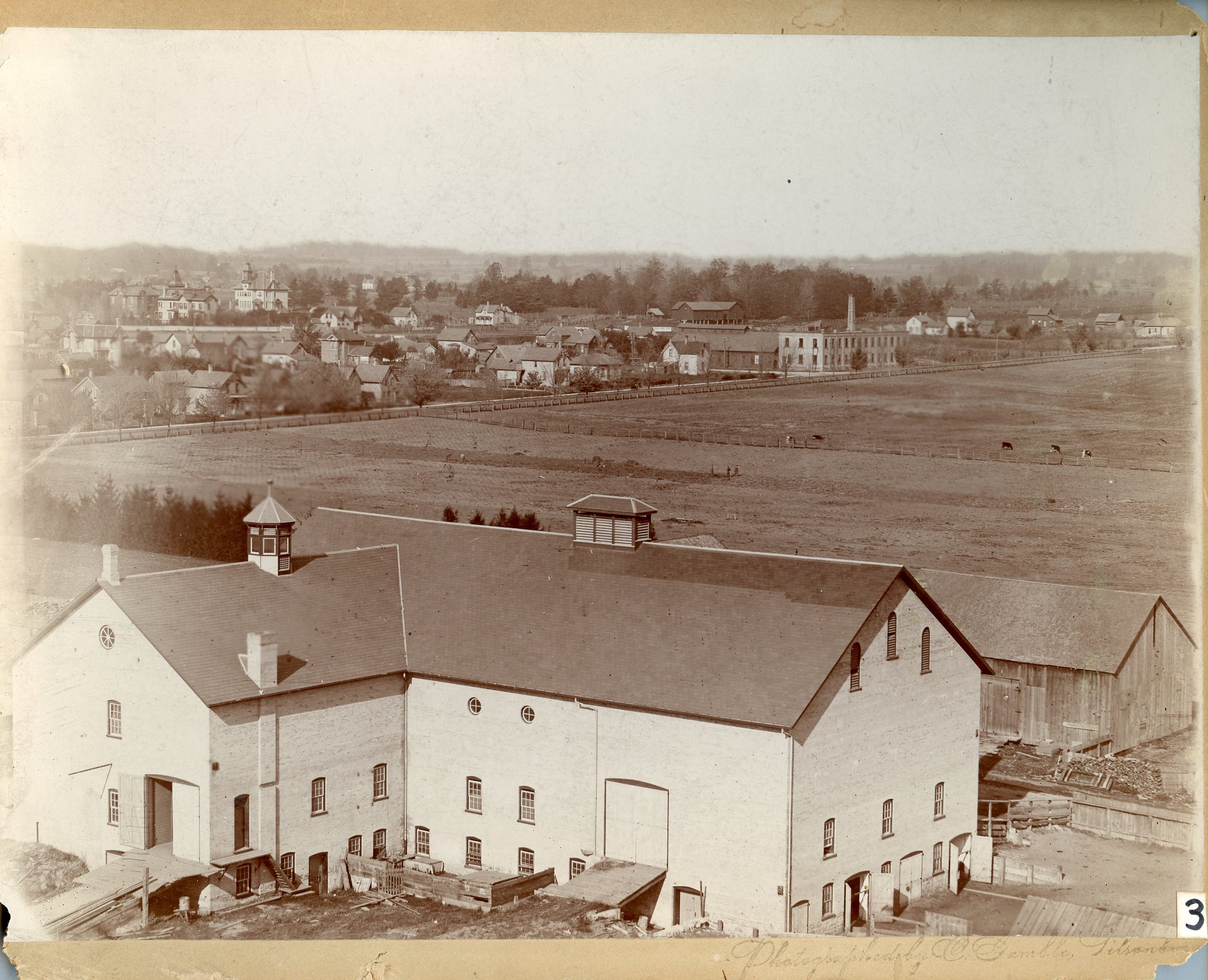 An aerial view of the Tillson's farm property. The town of Tillsonburg can be seen in the distance. In the foreground is a large white barn and a smaller frame building. Behind the barn is a large farm field with livestock grazing.